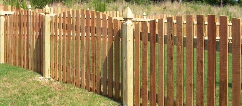 A new wooden board fence with a turned post cap, set on a green lawn under a clear sky.